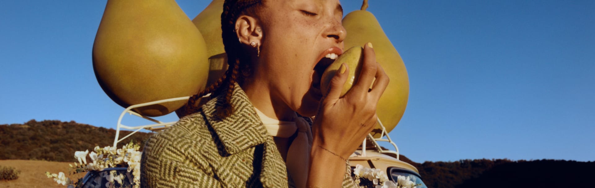 Adwoa Aboah with braided hair, biting into a pear with backdrop of three large pear props placed on top of a car roof rack.